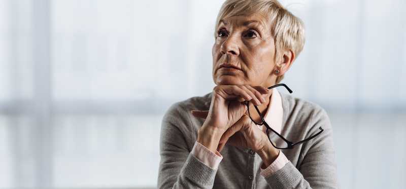 Person stares up thoughtfully while holding eyeglasses and sitting at desk.