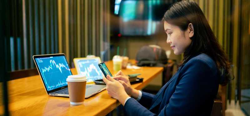Person looking at phone with stock market chart on laptop and paper cup nearby.