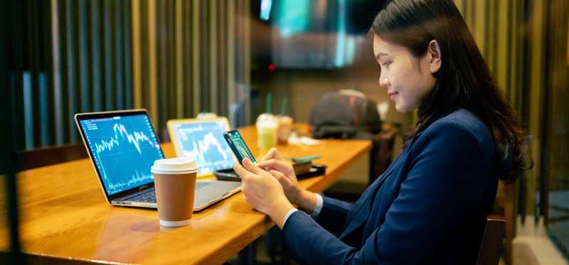 Person looking at phone with stock market chart on laptop and paper cup nearby.