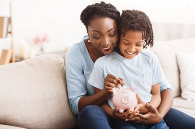 Adult holding child and putting coin in piggybank.