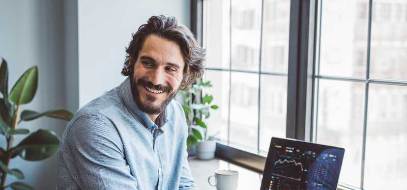 Smiling adult sitting at desk in front of window with laptop showing investing charts.