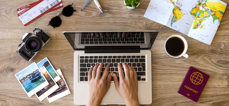 Hands of a person researching travel on a laptop that's sitting on a table with pictures, a passport, brochures, and other travel-related items.