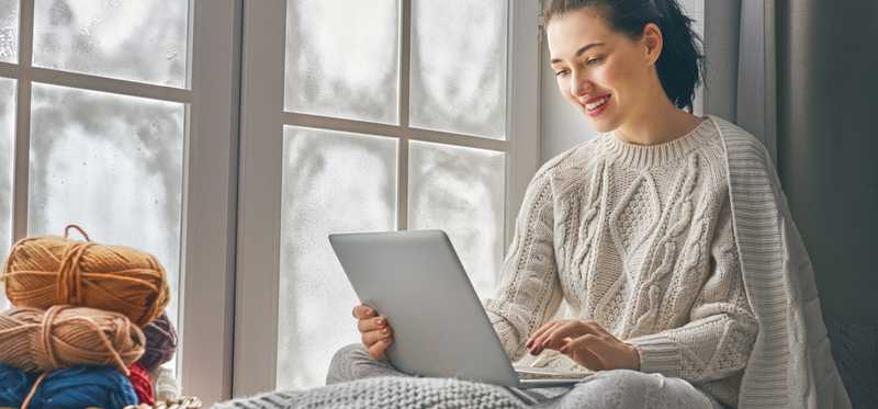 Woman in sweater smiling as she uses a laptop in her lap with yarn nearby