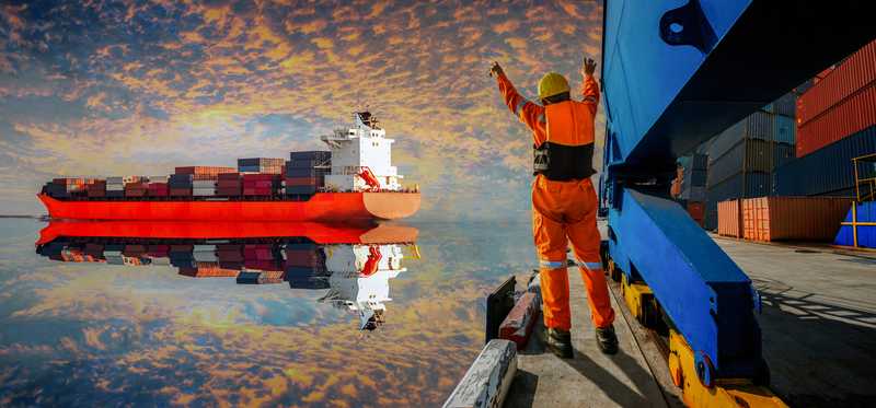 A person in orange signaling to a loaded freighter approaching a dock.