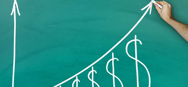 A hand is drawing a graph on a blackboard of an upward trending arrow, over a series of progressively bigger dollar signs.
