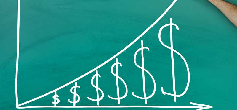 A hand is drawing a graph on a blackboard of an upward trending arrow, over a series of progressively bigger dollar signs.