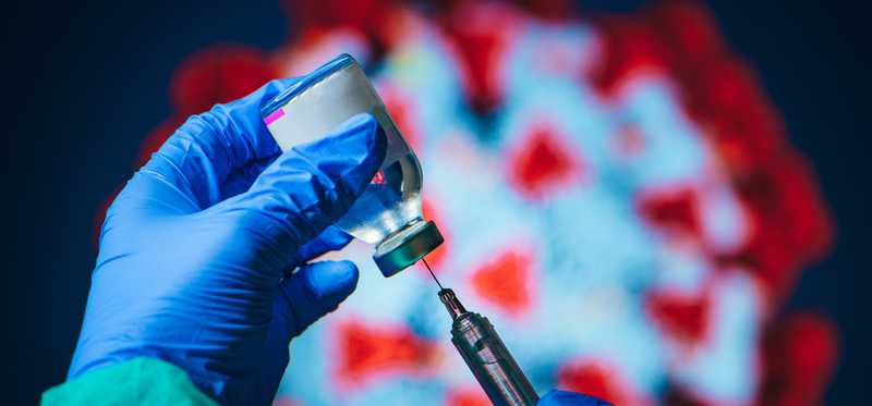 Gloved hands hold a vial and syringe with coronavirus illustration in background