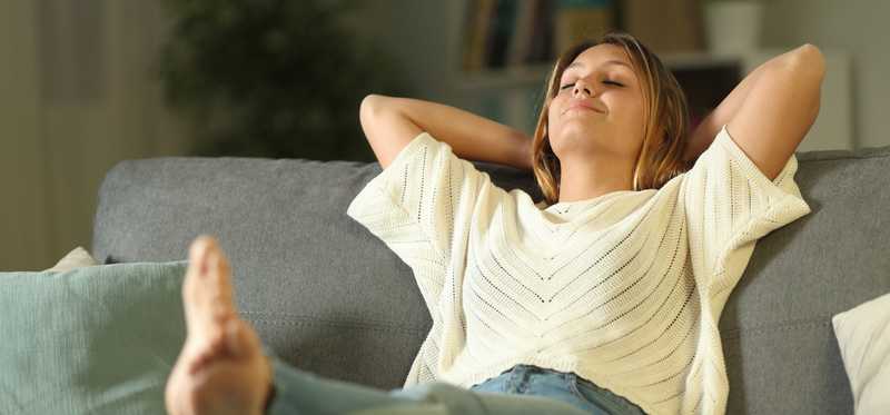 Smiling individual relaxing on couch, with eyes closed, arms behind their head, and their feet up on a table.