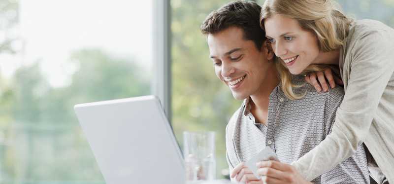 Two people smiling while looking at laptop near large window.