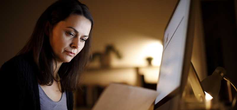 Adult in dark room looks thoughtfully at paperwork in front of laptop.