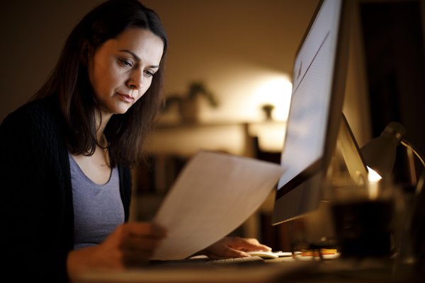 Adult in dark room looks thoughtfully at paperwork in front of laptop.