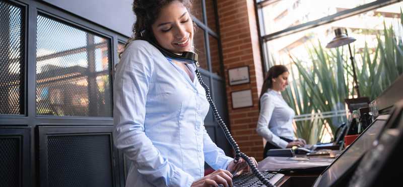 A receptionist answering the phone and typing on a computer at a hotel front desk.