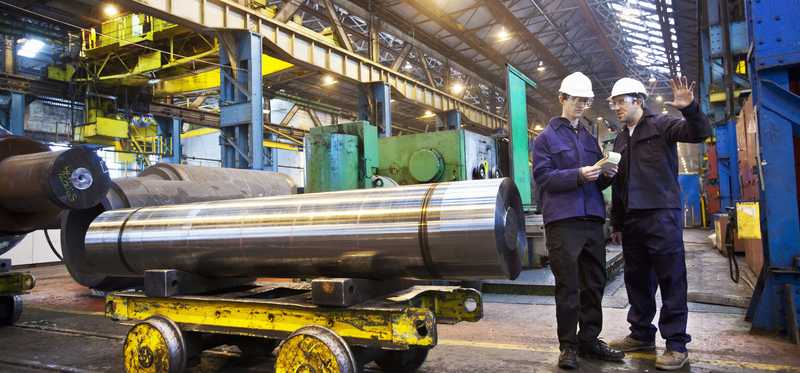 Two people talking while standing next to a steel roll inside a factory.