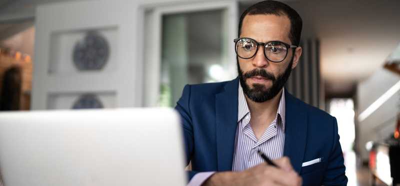 Adult wearing glasses takes notes thoughtfully while sitting at desk with laptop.