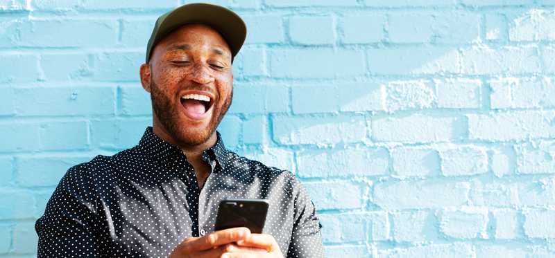 Individual wearing baseball cap smiles broadly while looking at smartphone screen outside.