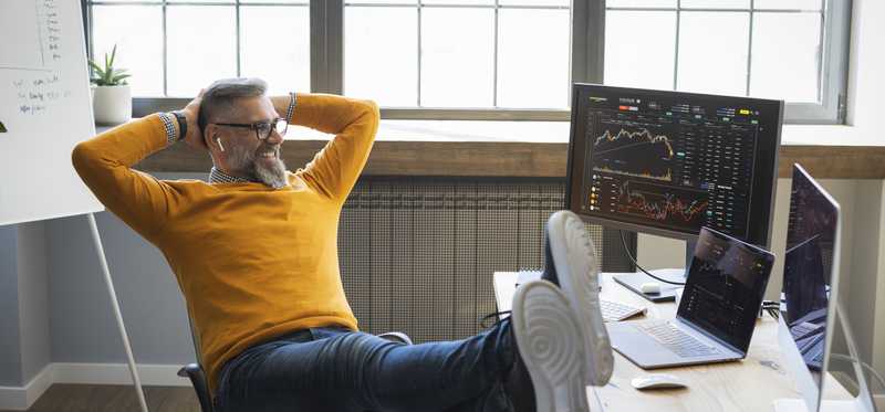 Person in orange sweater smiles while leaning back and putting feet on desk.