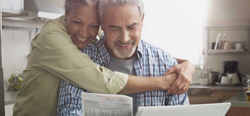 Person embraces another person while both smile and look at paperwork in kitchen.