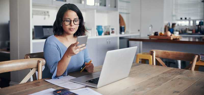 Person looks at phone while working on laptop in kitchen.
