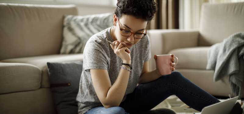 Person sitting on floor using laptop while holding coffee mug.