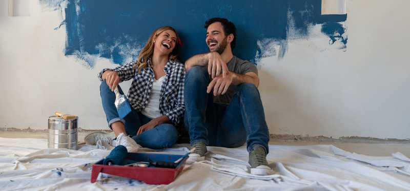 A couple is laughing as they sit in front of a white wall they are painting blue.