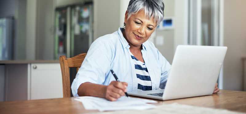 Person writes on paper next to laptop on kitchen table.