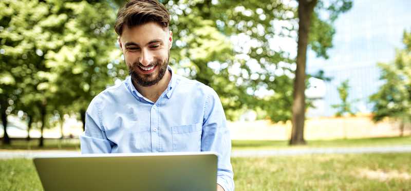Person looking at laptop and smiling while sitting on ground in park.