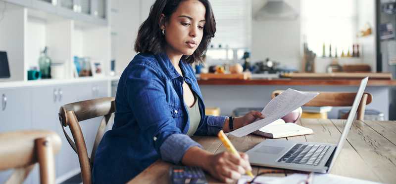 Person sitting at kitchen table and writing on paperwork near laptop.
