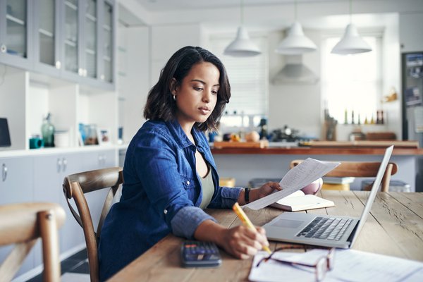 Person sitting at kitchen table and writing on paperwork near laptop.