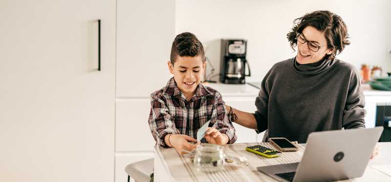 Parent smiles as child counts cash in jar at kitchen table.
