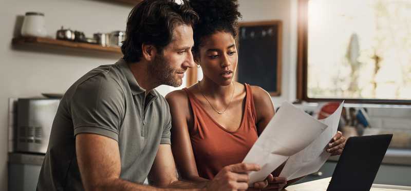 Two people study and compare paperwork at table with laptop.