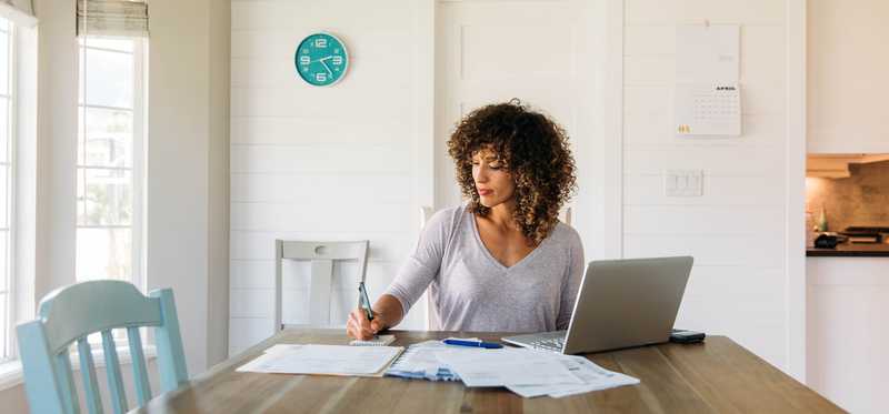Person sits at dining table with paperwork and laptop.