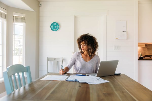 Person sits at dining table with paperwork and laptop.