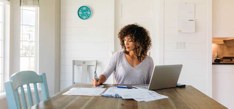 Person working at a laptop and writing on a notepad while sitting at a long wooden table.