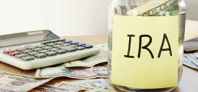 Jar of money labeled IRA sitting next to a calculator and atop various denominations of U.S. currency.