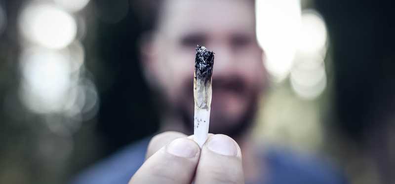 A bearded man holding a lit cannabis joint with his outstretched fingertips.