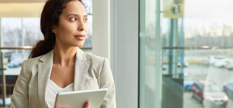 A businesswoman holding a tablet while looking out a window.