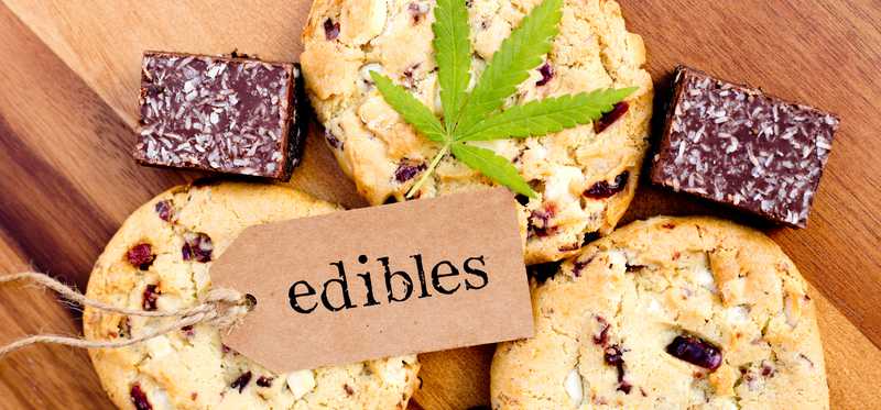 A cannabis leaf and a tag with the word edibles written on it that are lying atop an assortment of cookies and brownies.