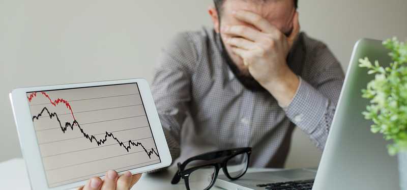 A man with one hand over his face holding up a plunging chart in the other.