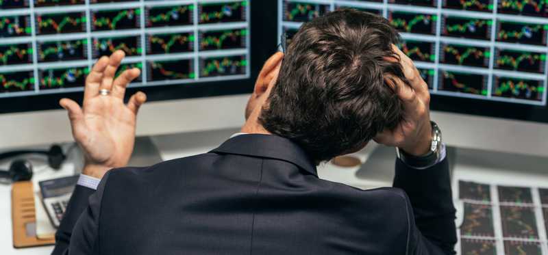A frustrated stock investor looking at multiple charts on their computer monitors and grasping the top of their head.