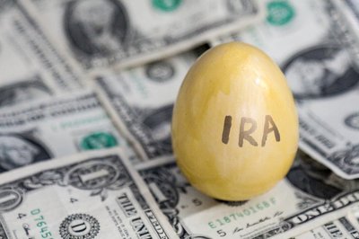 A golden egg with the word "IRA" written on it sitting on a messy bed of one dollar bills.