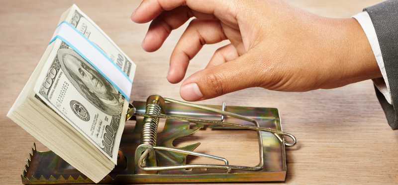 A hand reaching for a neat stack of hundred dollar bills in a mouse trap.