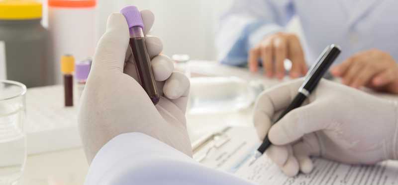 A lab researcher wearing gloves while examining a vial of blood and making notes.