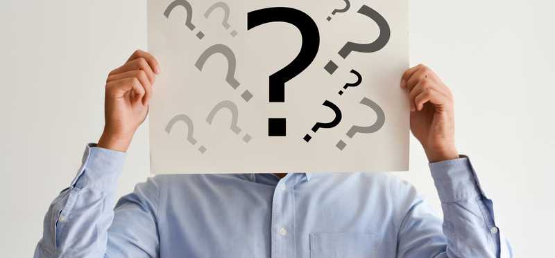 A man holding a cardboard sign in front of his face with numerous question marks drawn on it.
