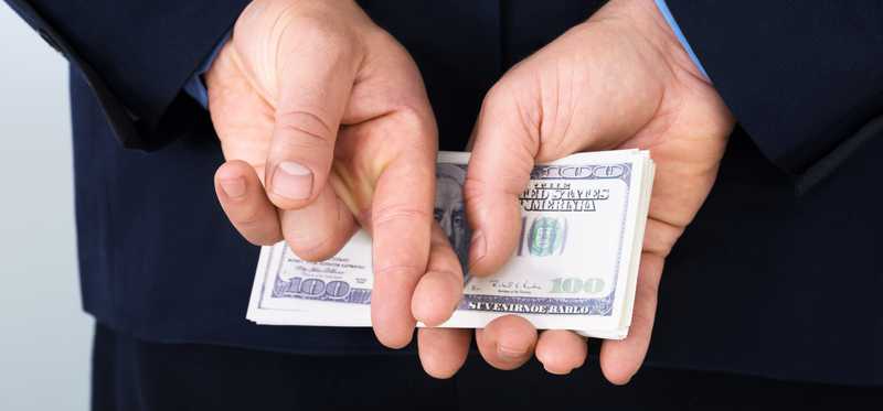 A man in a suit holding a neat stack of hundred dollar bills behind his back with his fingers crossed.