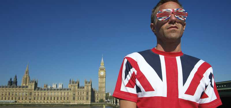 A man wearing a British flag print shirt and sunglasses standing in front of Parliament.