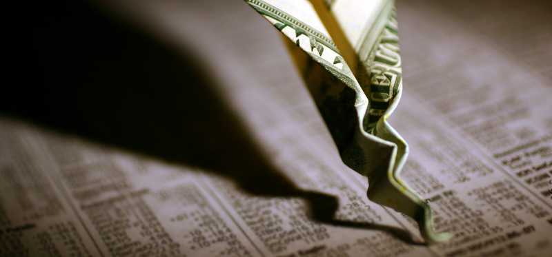 A paper airplane made out of a one dollar bill that's crashed and crumpled into the financial section of the newspaper.