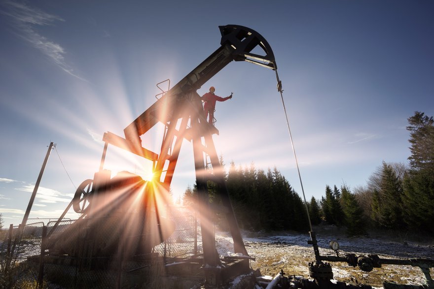 A person standing on an oil well with the sun shining in the background.