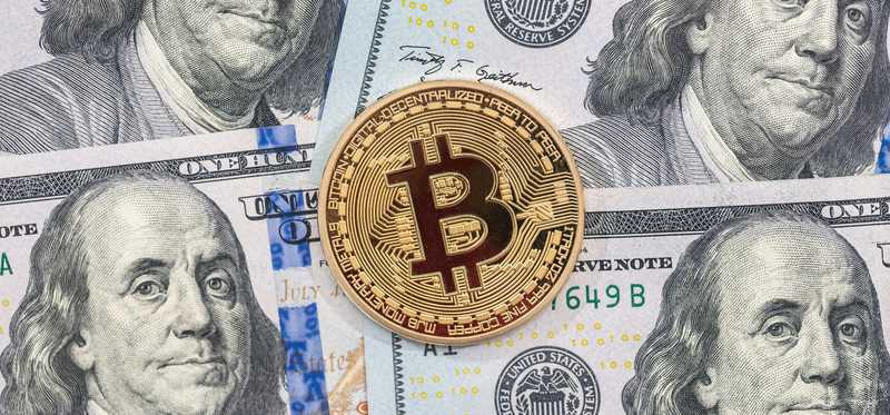 A physical gold coin marked with the bitcoin B lying atop hundred dollar bills.
