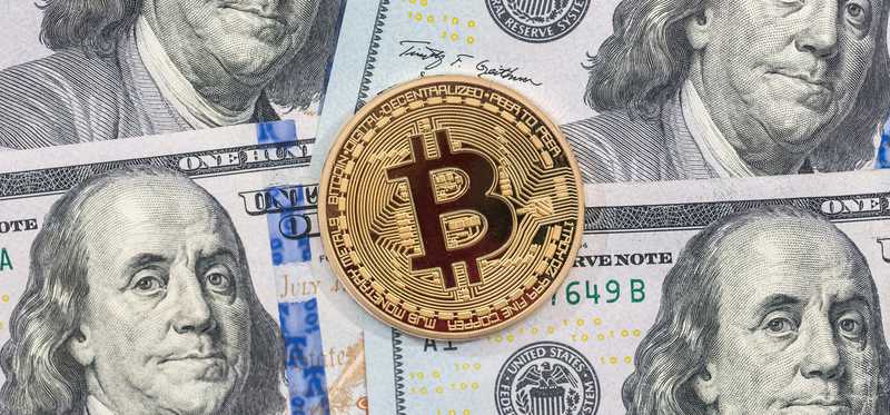 A physical gold coin marked with a B on top of a messy pile of cash.