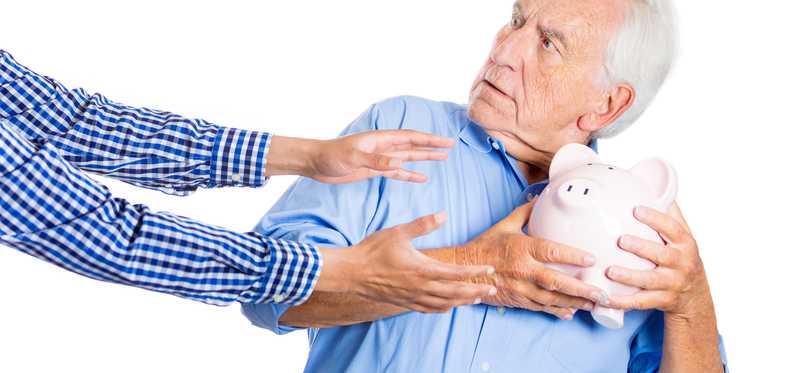 A senior man tightly clutching his piggy bank as outstretched hands reach for it.